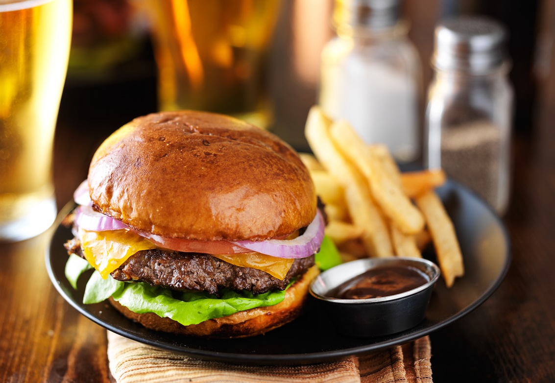 Close-up of a classic cheeseburger with lettuce, tomato, red onion, and cheddar cheese on a brioche bun, served with golden fries and dipping sauce on a black plate. A glass of beer is visible in the background.